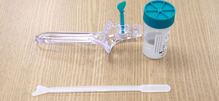 Espéculo vaginal descartável, frasco de coleta de amostra citológica e espátula cervical sobre superfície de madeira