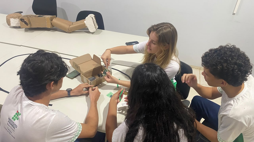 Estudantes reunidos em torno de uma mesa montando um protótipo de óculos com peças e caixas