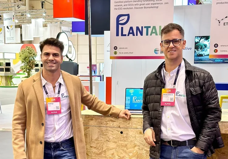 Dois representantes da Plantah em estande da empresa durante o Web Summit, com cartaz da marca e computador portátil sobre o balcão