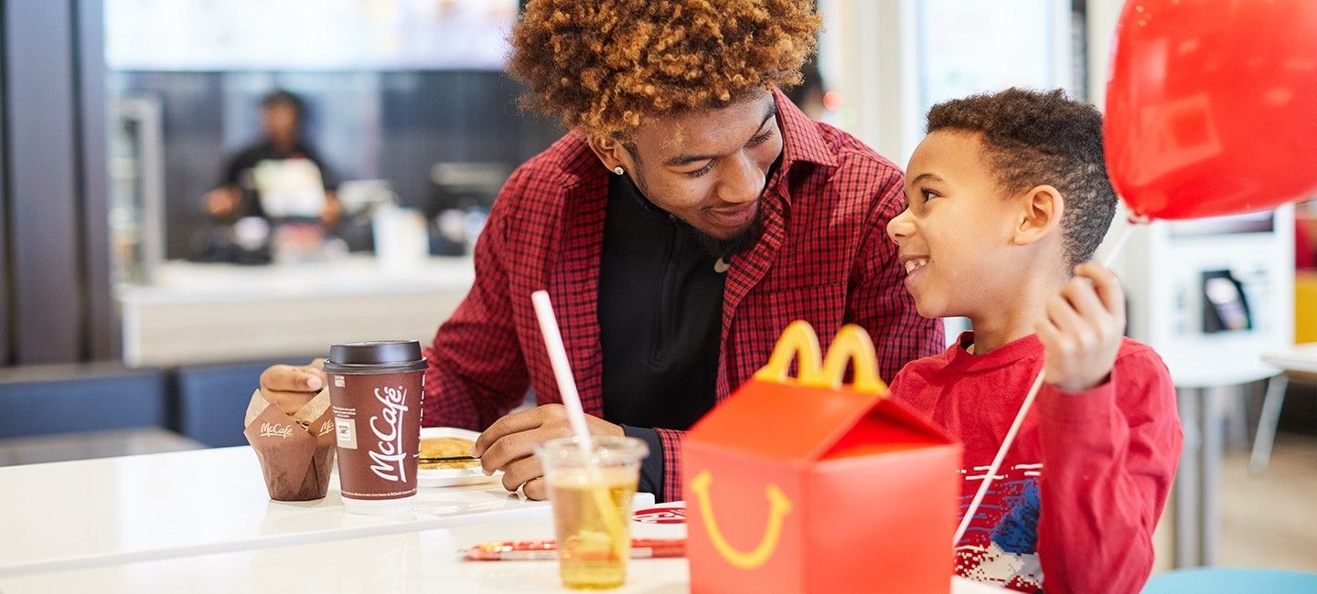 Pai e filho sorrindo em um restaurante McDonald’s, com lanche e balão vermelho sobre a mesa.