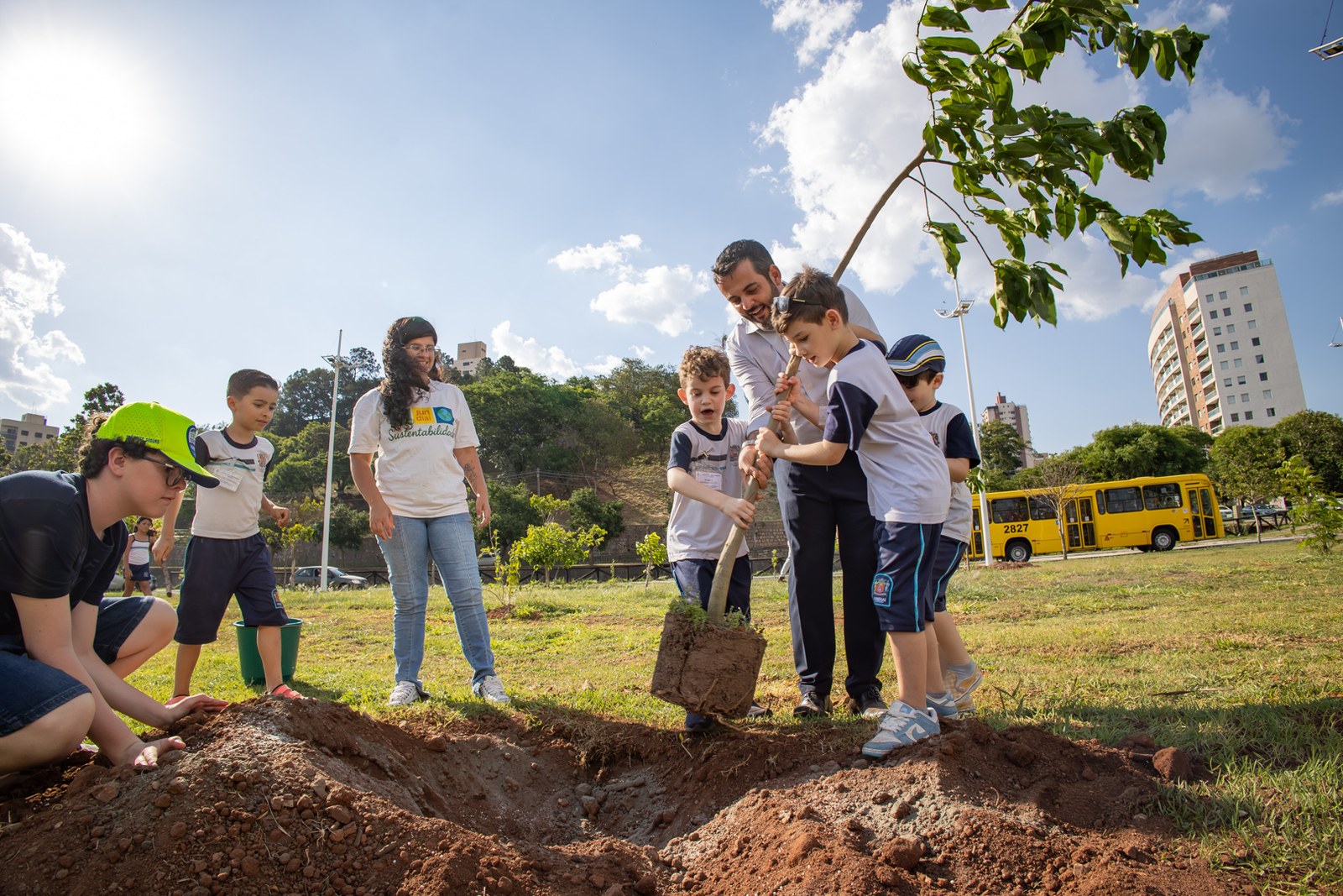 Estudantes uniformizados plantam uma árvore com apoio de educadores em área gramada